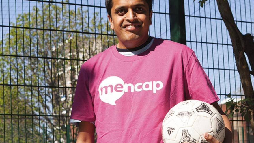 Vijay is standing outside next to a fence around a football pitch, wearing a pink Mencap t-shirt and holding a football. He is smiling at the camera,